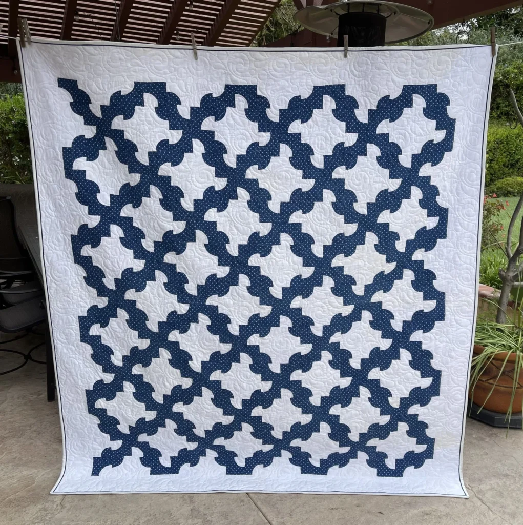 Mary-DP-quilt-vintage A large quilt with a white background and an intricate interlocking blue geometric pattern, displayed outdoors on a patio.