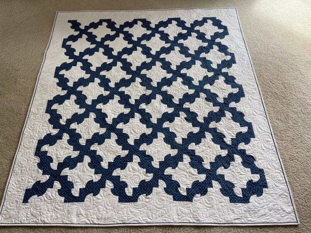 Mary-DP-showing-quilting A quilt featuring a repeating blue geometric pattern on a white background, with intricate stitching details on the white areas, is laid flat on a beige carpet.