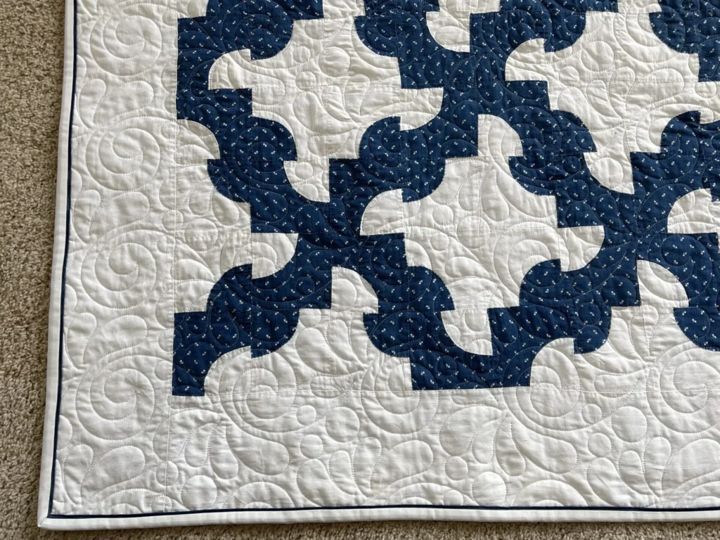 Mary-corner-DP-quilt A close-up of a quilt corner with a navy blue and white geometric pattern and intricate swirl quilting, displayed on a beige carpet.