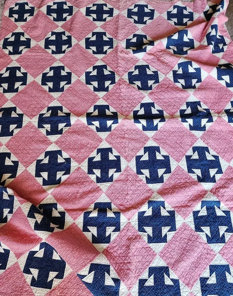 A quilt featuring a repeating pattern of navy blue and white geometric shapes on a pink background, forming a grid design with diamond-shaped blocks. The quilt appears slightly wrinkled.