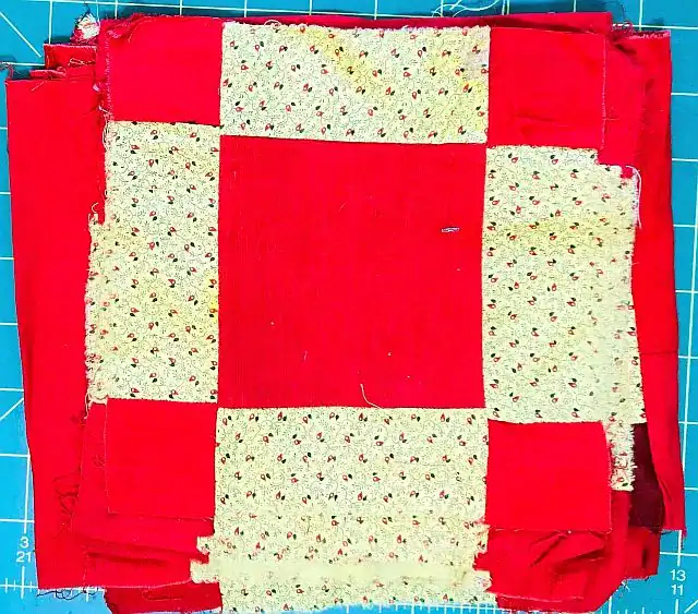 A stack of quilt blocks made of red and cream patterned fabric arranged in a square-in-a-square design, placed on a teal cutting mat with gridlines.