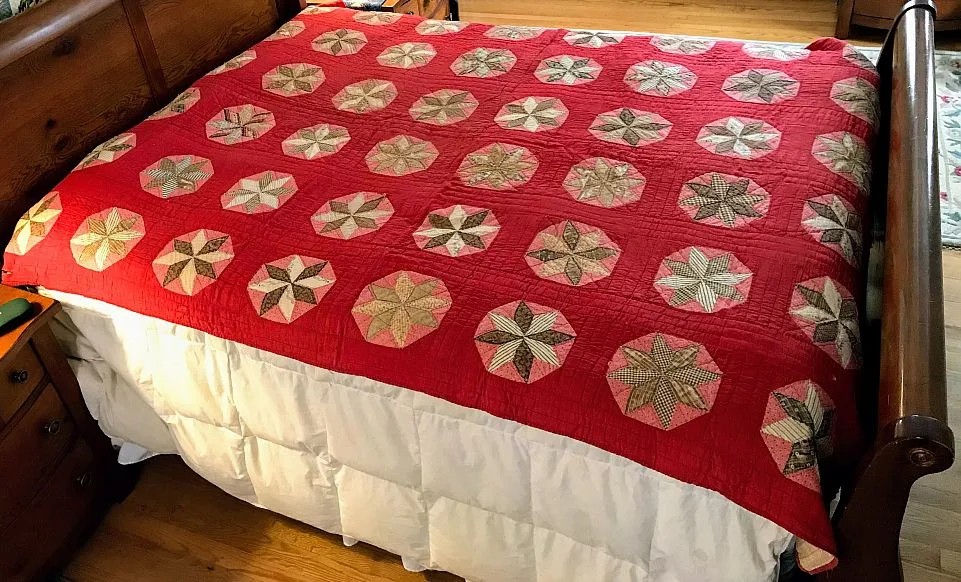 A wooden bed is covered with a red quilt featuring a pattern of circular, star-like designs in shades of beige, green, and brown. White bedding is visible underneath. The bed is in a warmly lit room with wood floors.