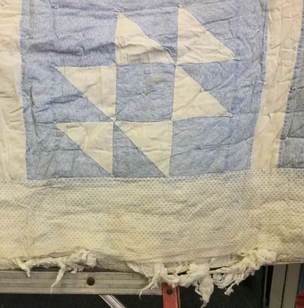 A worn quilt with a blue and white geometric pattern and a frayed edge, showing visible signs of age and use. The quilt rests on a metal surface.