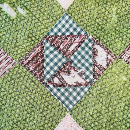 A quilt block with a green checkered diamond center, brown-and-white marbled triangles, and a green floral background. The quilt is covered with a clear protective layer.