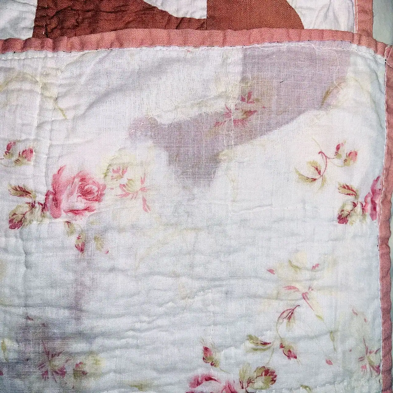 A close-up of a quilt with a faded floral pattern, mainly featuring pink roses and green leaves on a white background, bordered by light pink fabric.
