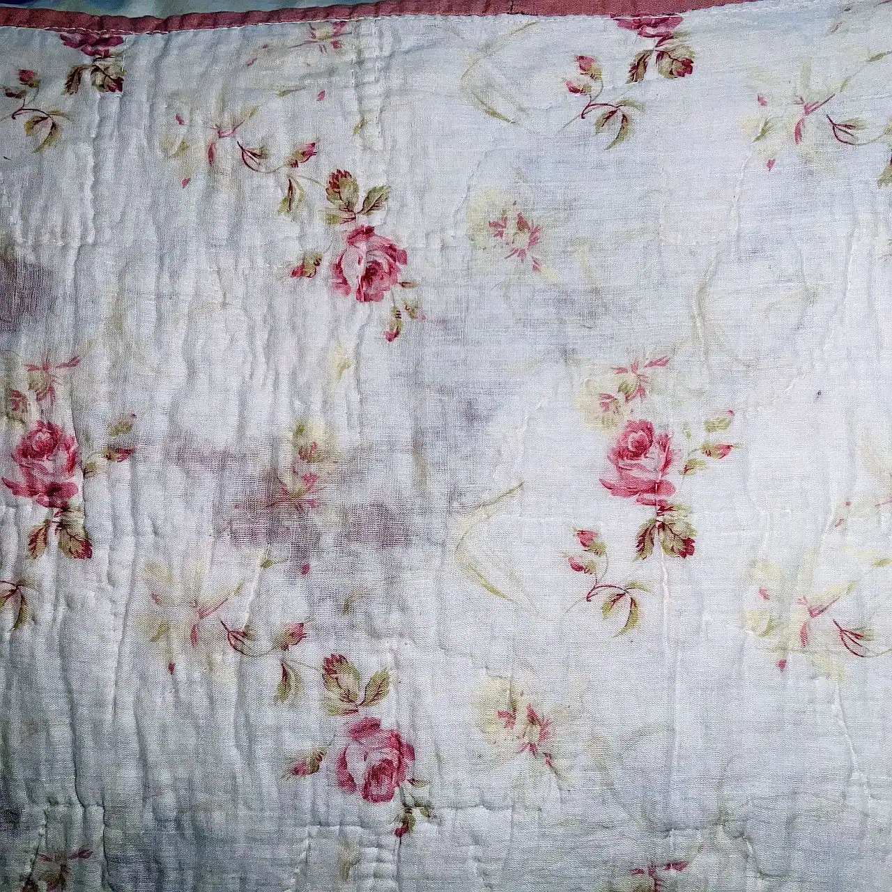 A close-up of a fabric with a faded floral pattern featuring pink roses and green leaves on a white background. The fabric appears old and wrinkled.