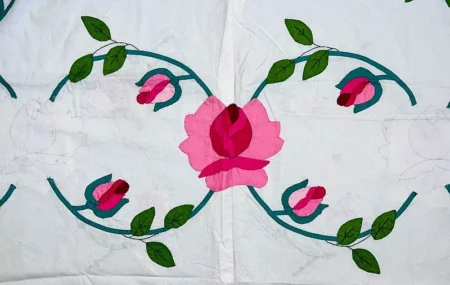 A quilt section with appliqué roses and rosebuds in shades of pink, green leaves, and teal stems on a white background. Drawing inspiration from quilt history, some floral outlines remain unfinished, adding a touch of timeless charm to the classic design. For more details or inquiries, please visit our About and Contact pages.