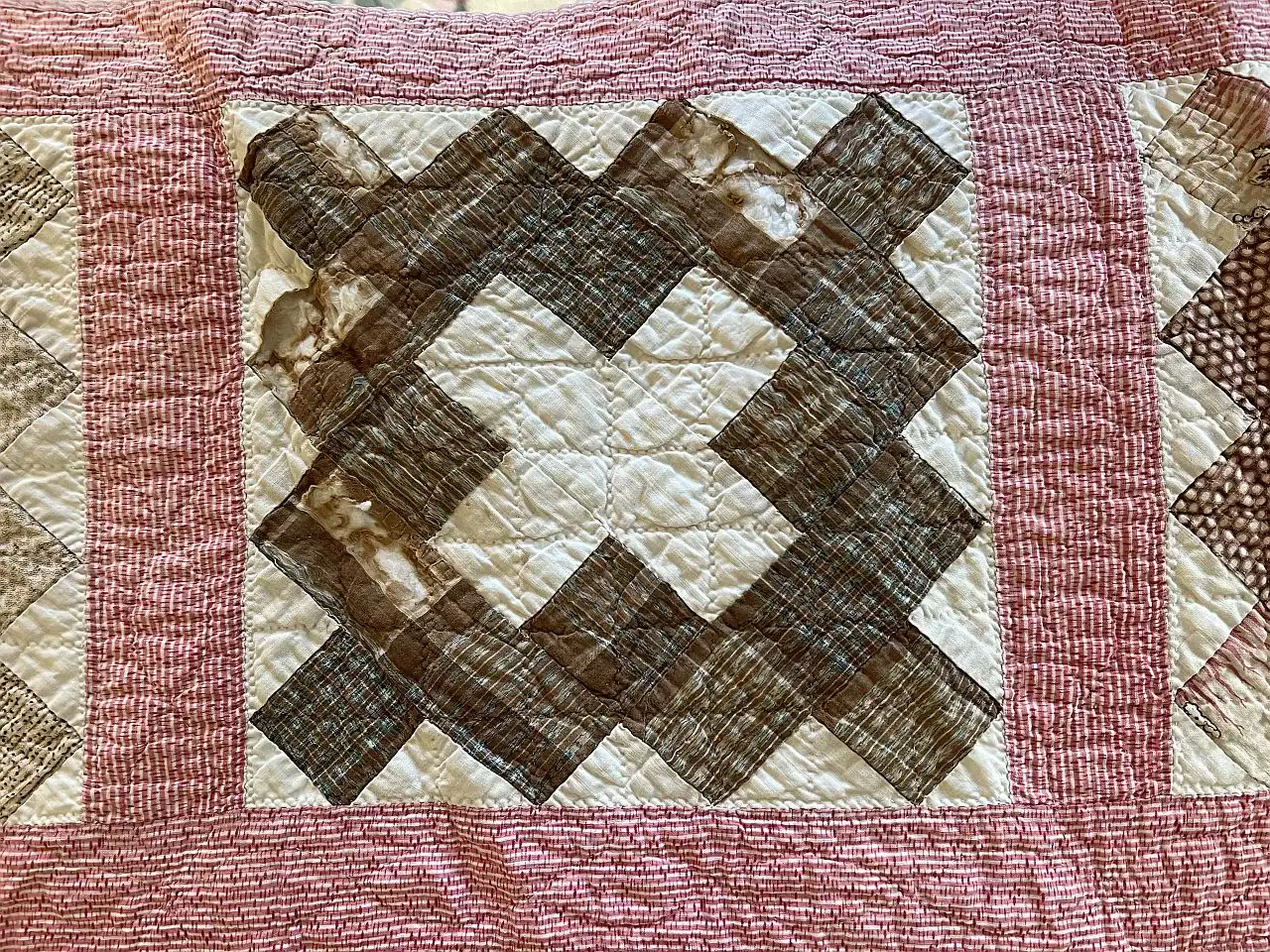 A quilt block with brown and cream geometric patterns arranged in a diamond shape, bordered by pink and white striped fabric. The quilt features visible stitching in a crisscross pattern.