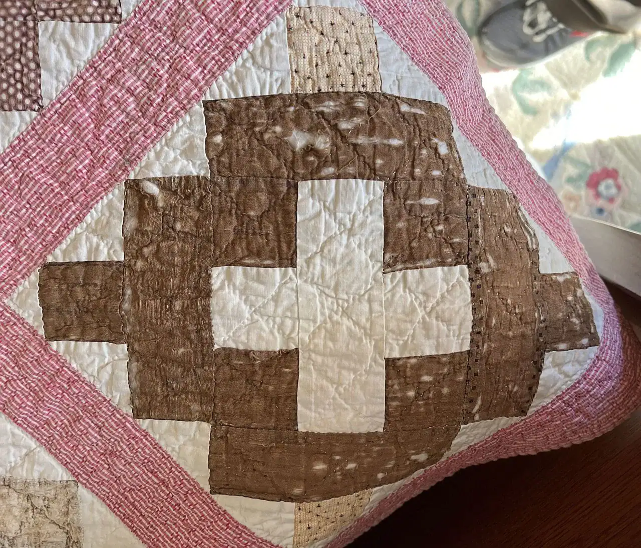 A close-up of a quilt corner with a brown and white cross pattern, bordered by pink and white strips. Part of a shoe and floral rug are visible in the background.