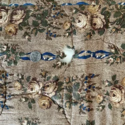 A quarter lies on wrinkled, floral-patterned fabric with a burn hole in the center, showing white stuffing beneath. The fabric has beige roses, green leaves, and a blue ribbon design.