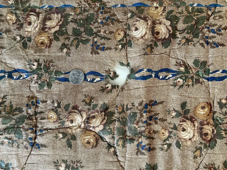 A quarter lies on wrinkled, floral-patterned fabric with a burn hole in the center, showing white stuffing beneath. The fabric has beige roses, green leaves, and a blue ribbon design.