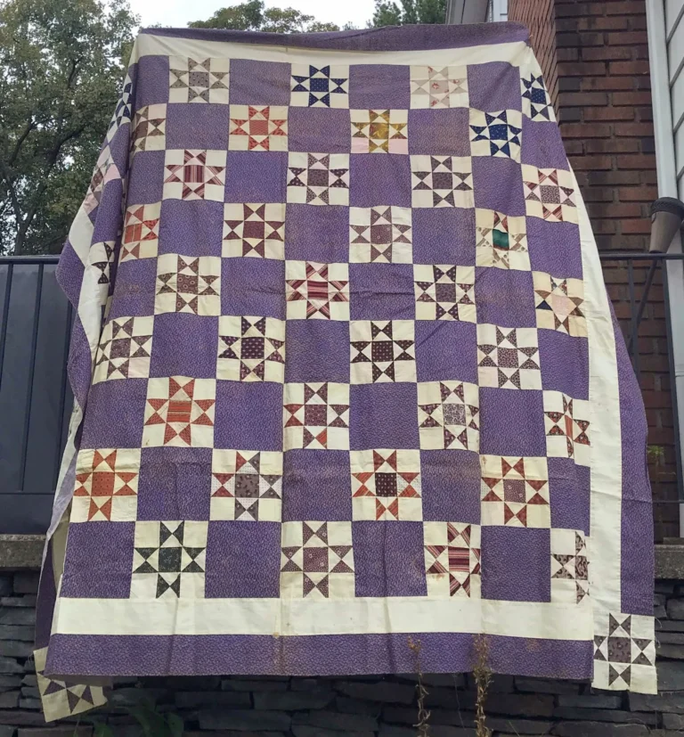 A large patchwork quilt from Telltale Textiles: Double Purple, featuring purple, white, and multicolored pinwheel star patterns, hangs outdoors against a brick house and railing.