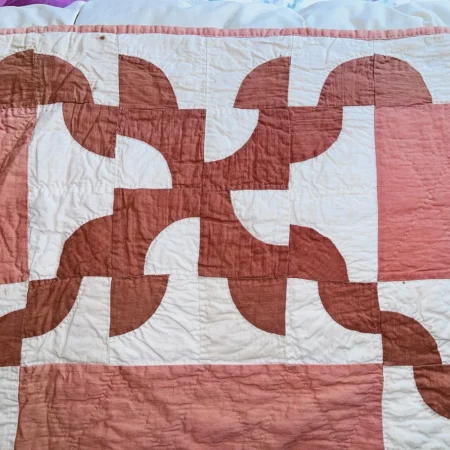 A quilt with a geometric pattern in pink and white, featuring curved shapes and intersecting lines forming a repeating abstract design.