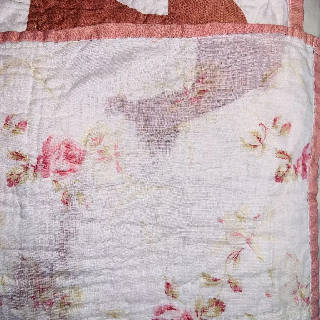 A close-up of a quilt shows faded, wrinkled white fabric with a pink floral pattern and pink stitching along the seams. A brown patch appears at the top, and the quilted stitches are visible throughout.