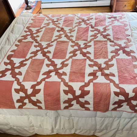 A bed with a quilt featuring a pink and white geometric pattern of squares and interlocking vine shapes, spread out neatly on a white comforter.