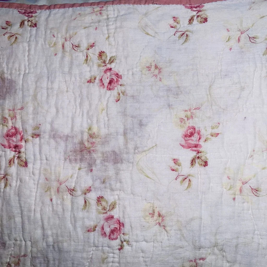A close-up of a white quilt or fabric with a pink rose floral pattern and subtle green leaves. The fabric appears slightly wrinkled and has a worn, vintage look.