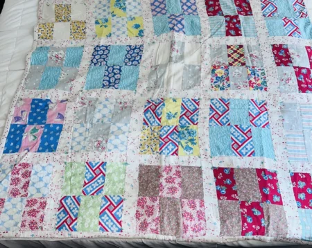 A handmade quilt with a patchwork pattern, featuring colorful squares of floral, geometric, and solid fabrics in blue, red, pink, and white tones, spread across a bed.