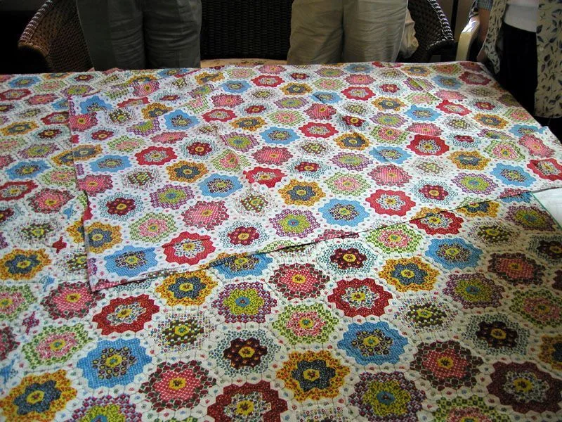 A colorful patchwork quilt with flower-like hexagon patterns in various bright colors is spread on a table. Several people stand around the table, with only their lower bodies visible.