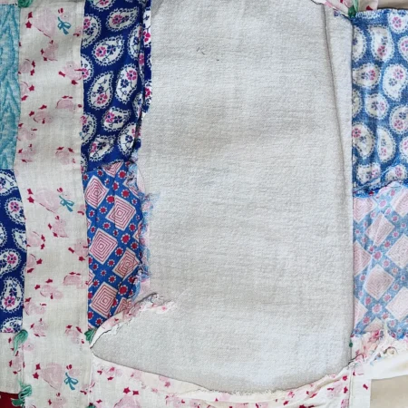 A patchwork quilt with faded, worn fabric pieces in various pastel and blue patterns, including florals, paisleys, and geometric prints. The center area is a plain, light-colored fabric.