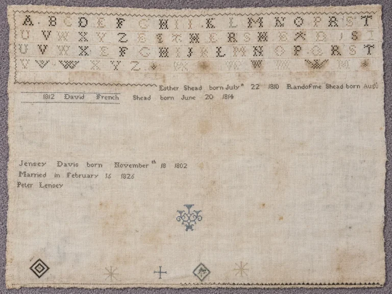 A vintage sampler features rows of stitched alphabets, names, dates of birth, a marriage date, and decorative motifs on faded fabric with a stitched border.
