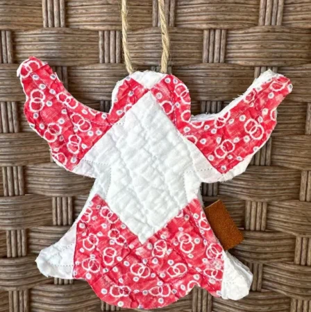 A quilted angel ornament with red and white fabric hangs from a twine loop, showcasing the artistry often celebrated by those who repair, restore, or conserve quilts. The background is a woven wicker surface.