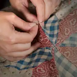Close-up of hands sewing fabric pieces with blue, red, and tan patterns, creating a patchwork quilt by hand with a needle and thread.
