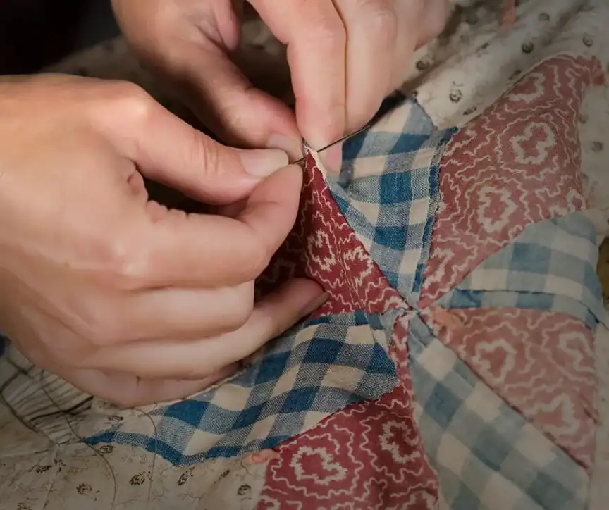 Close-up of hands sewing fabric pieces with blue, red, and tan patterns, creating a patchwork quilt by hand with a needle and thread.