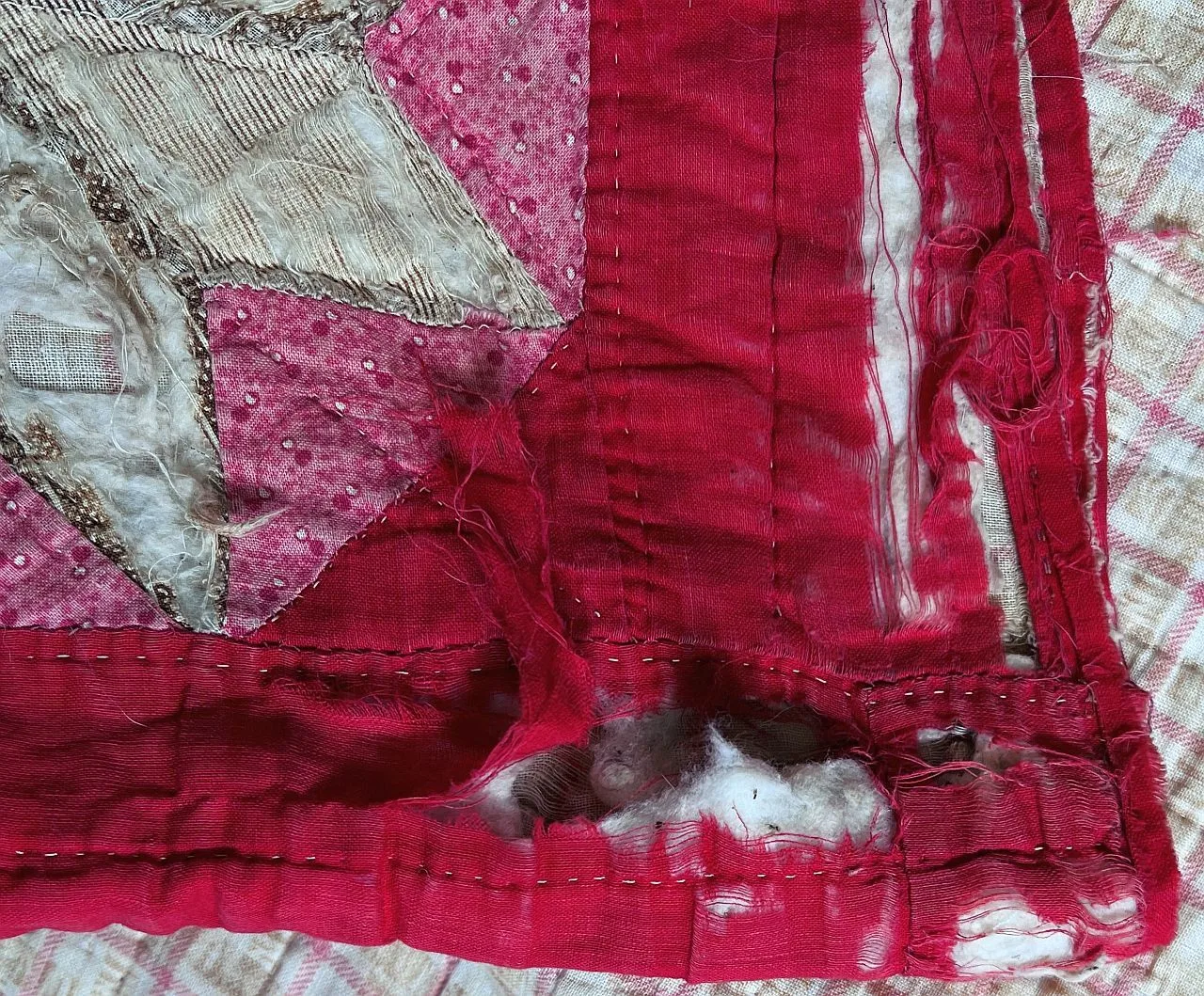 Close-up of a worn quilt corner with red and pink fabrics, fraying edges, and visible stitches. Some fabric is torn, revealing batting underneath. Patterns include dots and stripes. The quilt appears old and well-used.