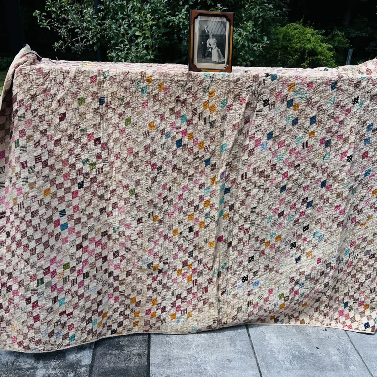 A large quilt made of small, colorful diamond-shaped pieces is draped outdoors over a railing. A framed black-and-white photograph rests on top. Trees and greenery are visible in the background.