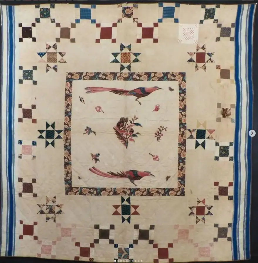 A quilt with a large central square featuring two birds and flowers, surrounded by patterned borders and multicolored patchwork stars and squares, with stripes along the sides.