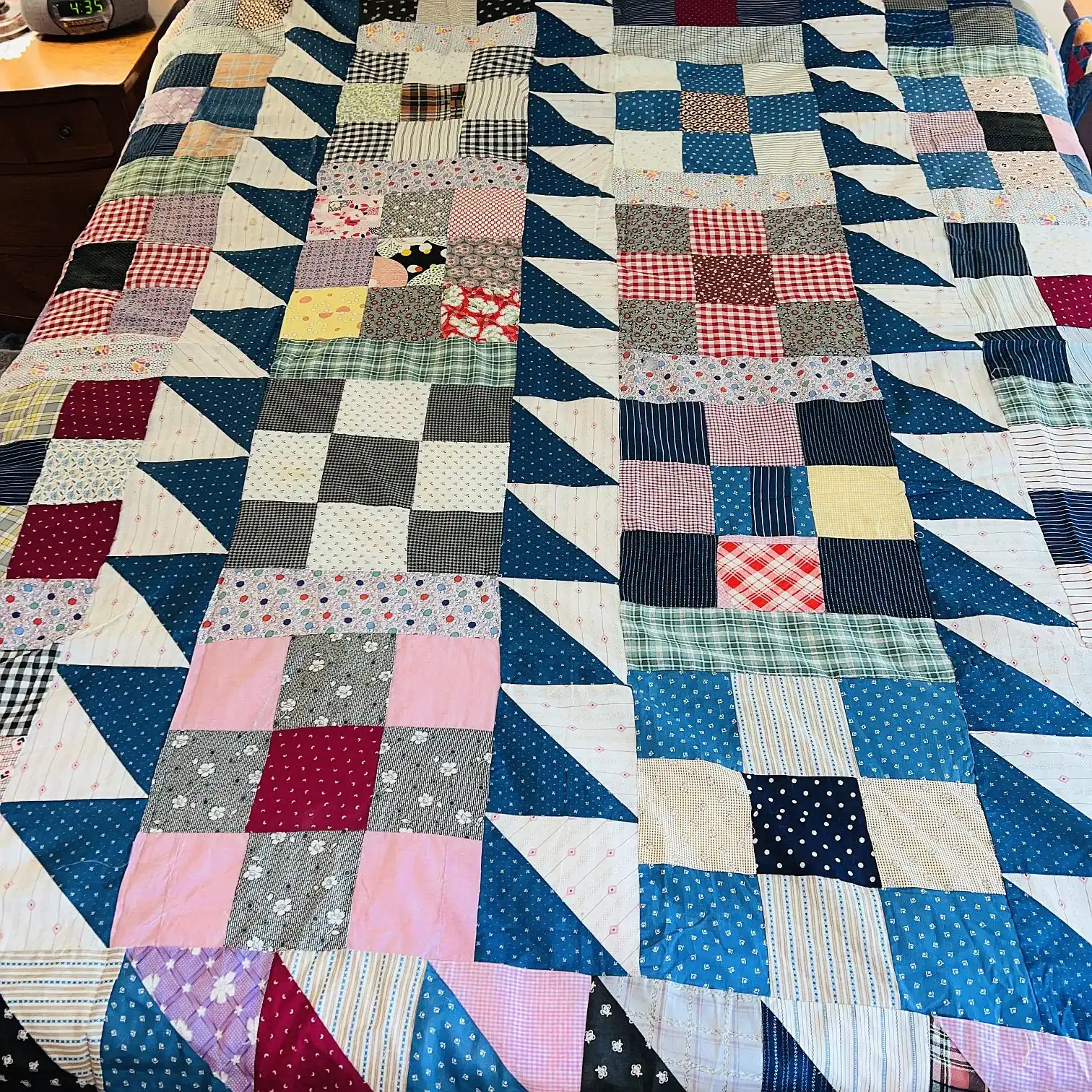 A colorful patchwork quilt covers a bed, featuring squares and triangles in various patterns and colors, including blue, pink, red, plaid, and polka dots. The quilt has a geometric design.