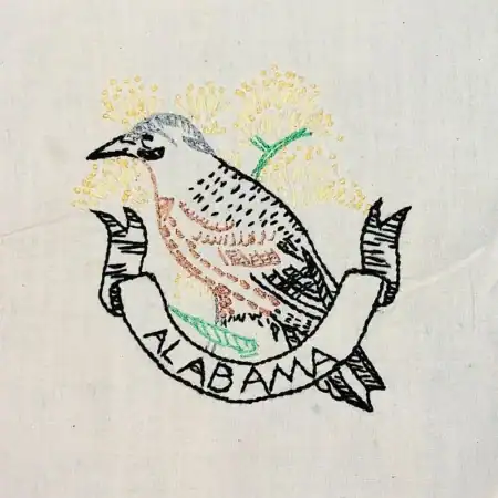 Alabama-flicker-on-goldenrod Embroidered image of a bird perched above a ribbon with the word Alabama, with yellow flowers in the background, all stitched on white fabric.