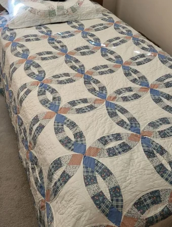 Arch DWR A bed covered with a handmade quilt featuring an interlocking ring pattern in blue, white, and pink fabrics. A matching pillow rests at the head of the bed. The quilt has a vintage, patchwork design.