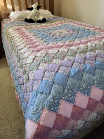 A neatly made bed with a colorful patchwork quilt in pastels and florals. A black and white stuffed cow sits atop a white pillow at the head of the bed.