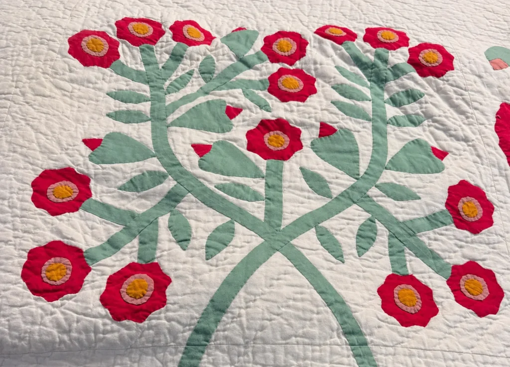 A quilt with green stems and leaves forming a symmetrical pattern, topped with red flowers that have yellow centers, on a white background.