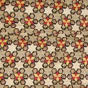 A patterned fabric with repeating hexagons, each containing floral designs and red, yellow, and black geometric shapes on a beige background. Reminiscent of classic cheater cloth faux patchwork, the pattern creates a visually intricate, vintage look.