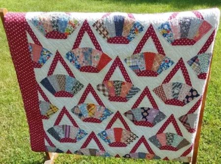 A quilt with a fan pattern made of multicolored, patterned fabric pieces, arranged in rows and framed by a red and white polka dot border, is draped over a wooden rack on green grass.