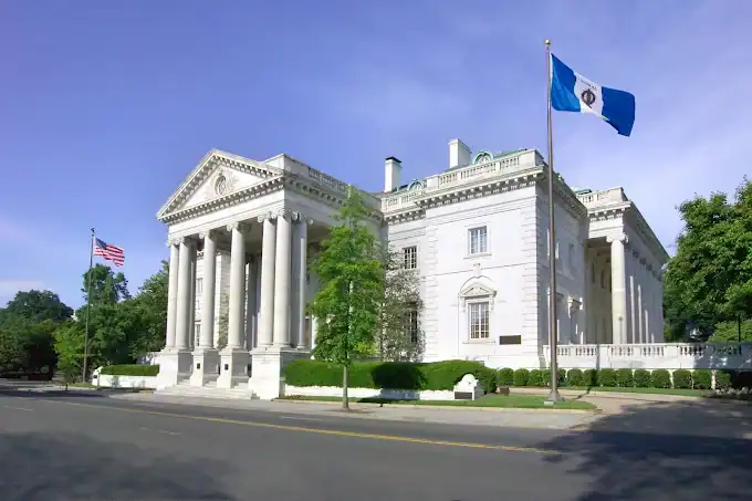 DAR Building A large white neoclassical building with columns, two flags flying on poles outside—one American, one blue and white—under a clear blue sky, with trees lining the street in front.