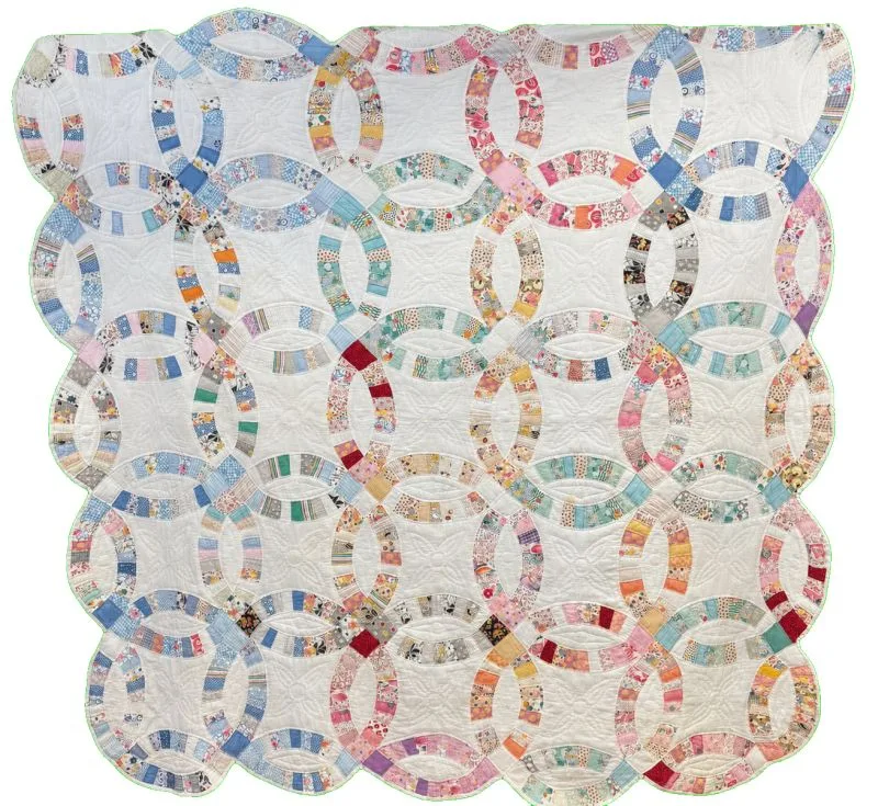 A handmade quilt with a double wedding ring pattern, featuring interlocking rings of multicolored fabric pieces on a white background. The fabric pieces are pastel and patterned, creating a patchwork effect.