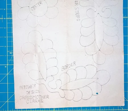 A quilt pattern sheet with sketched feather designs, labeled CENTER, BORDER, and FOR STRIPS. Text at the bottom reads FEATHER DESIGN GRANDMOTHER CLARKS. The background features a blue cutting mat—an inspiring example of vintage motifs found in Grandmother Clark Quilt Booklets.