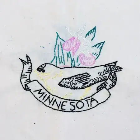 Minnesota-Goldfinch Embroidery of a bird lying on its back atop a banner reading MINNESOTA, with two pink flowers and green leaves in the background, stitched on white fabric.