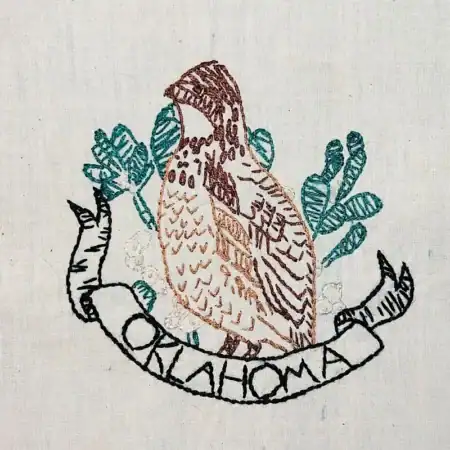 Oklahoma-Bobwhite Embroidery of a brown bird with green leaves behind it and a ribbon below reading OKLAHOMA on white fabric.