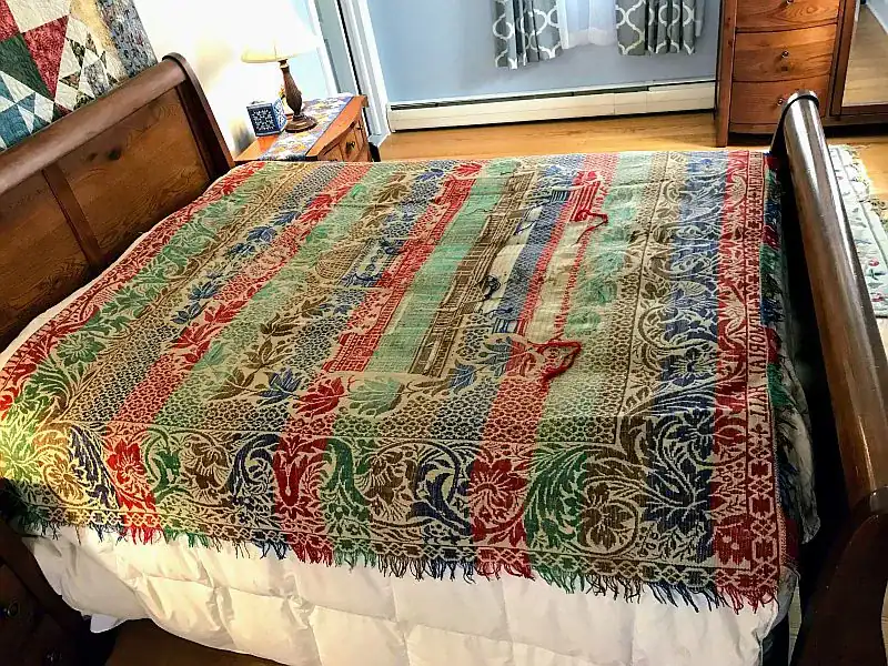 Philadelphia-Exposition-Coverlet A colorful, patterned woven blanket with red, green, blue, and gold designs is spread neatly over a wooden bed in a cozy, well-lit bedroom with a nightstand, lamp, and dresser visible.