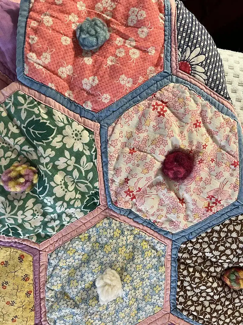 Potholder-Style Quilt A close-up of a colorful quilt with hexagonal patches in various floral patterns, each featuring a small, textured tuft or knot in the center.