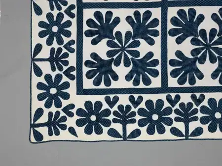 Quilt,_Oak_Leaf_pattern_1860-MET A section of a white quilt with navy blue stitched flower and leaf patterns along the border and inside squares, displayed on a plain gray surface.