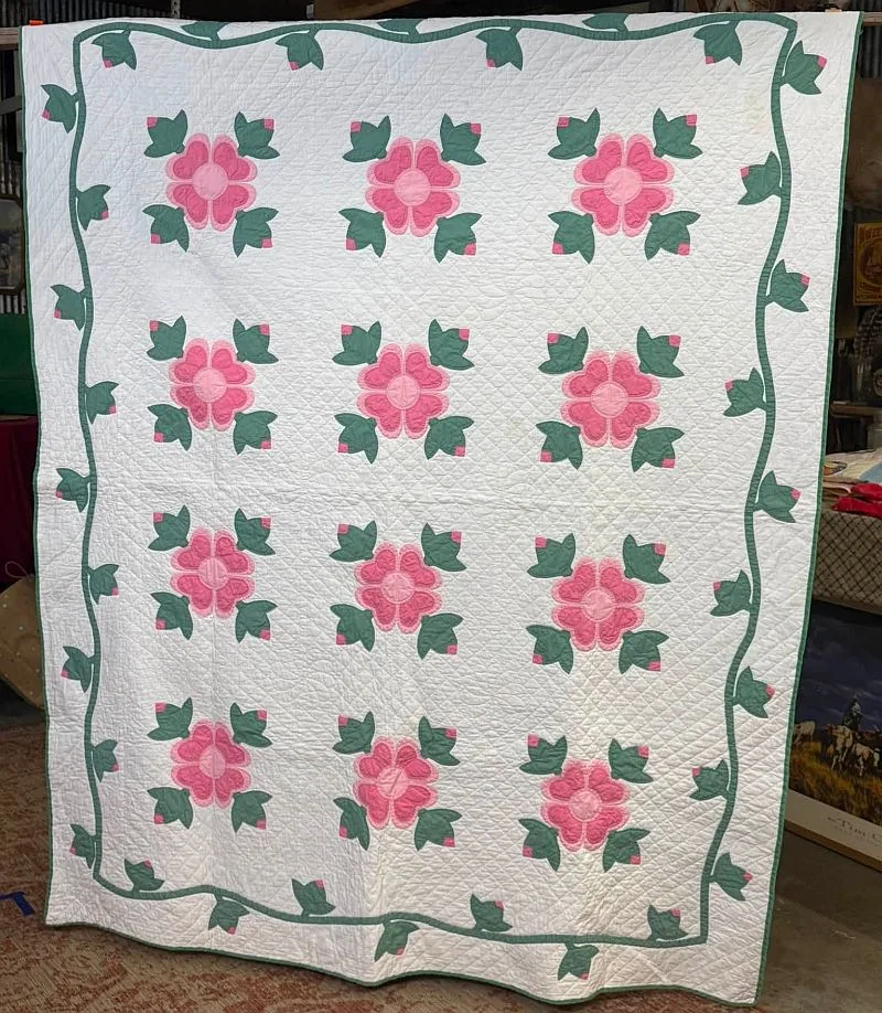 A handmade quilt with a white background features a repeating pink and green floral pattern and a green vine border. Reminiscent of classic designs highlighted in the February 1997 QHL digest, the quilt hangs vertically, showing its detailed stitching and garden-inspired design.
