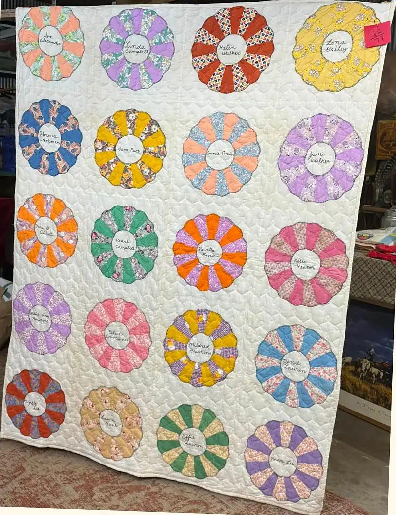 A handmade quilt with colorful circular flower patterns, each featuring a name written in the center, hangs on display indoors among various objects.