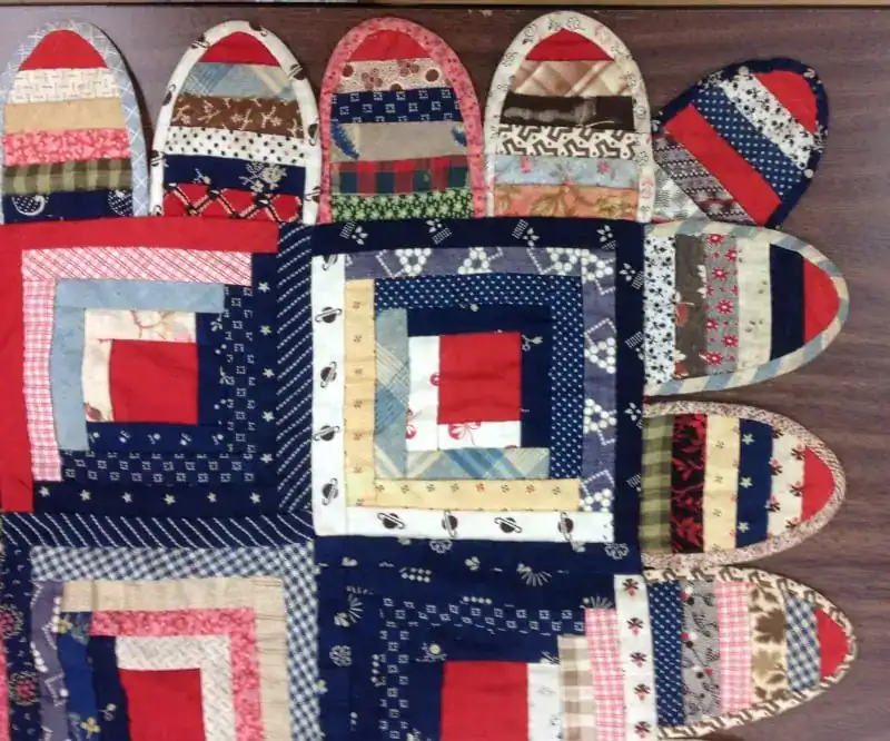 A handmade quilt with multicolored rectangular patches and semi-oval shapes along one edge, featuring various patterns in red, blue, beige, and white fabrics, displayed on a wooden surface.