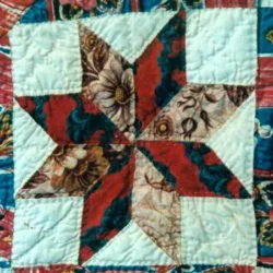 A quilt block featuring a star pattern with floral and red fabric pieces, bordered by light blue patches, displaying detailed stitching and intricate fabric designs—an excellent example for those interested in Dating Vintage Quilt Blocks.