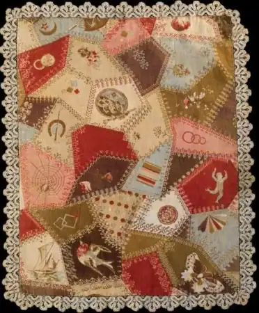 A colorful, patchwork quilt with embroidered designs and a lacy border, this piece features the charming look of Cheater Cloth Faux Patchwork. Various geometric shapes and images, like flowers, animals, and fans, decorate the quilt. The edges are trimmed with detailed lace.